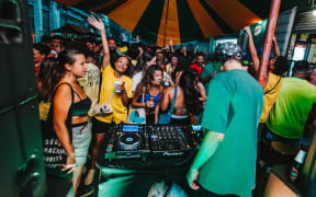 A group of colourfully dressed people surround a DJ on his decks.