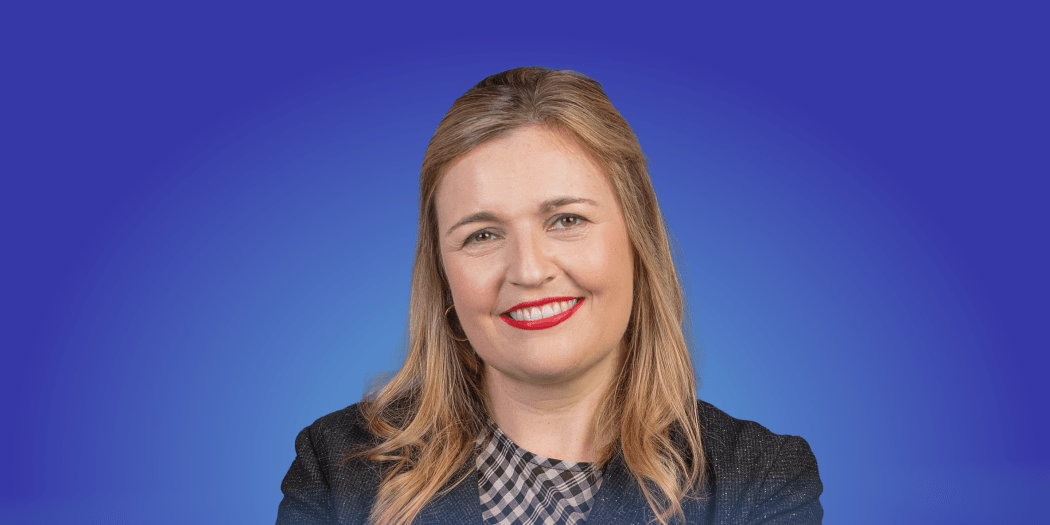 Charlotte Ryan smiles wearing a dark grey jacket and bright red lipstick