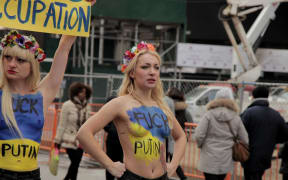 a woman standing amid a protest, bare-chested, with body paint saying "fuck putin"
