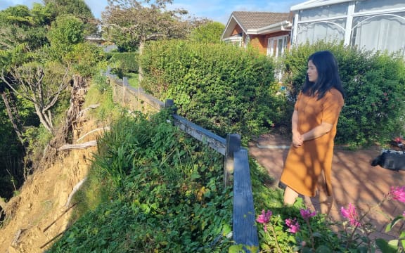 Jennifer Chen has black long hair and wears an orange dress. She stands at the edge of her property looking down at the cliff which has fallen away.