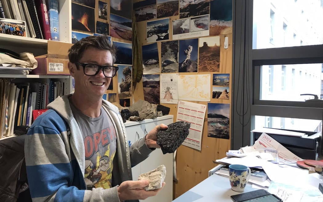 A man with glasses wearing a graphic tee and a blue and grey stripey hoodie sits behind a desk. He is smiling at the camera and holding two rocks, one black and one white. Behind him, the walls are covered with images of volcanoes and books are stacked in a bookcase.