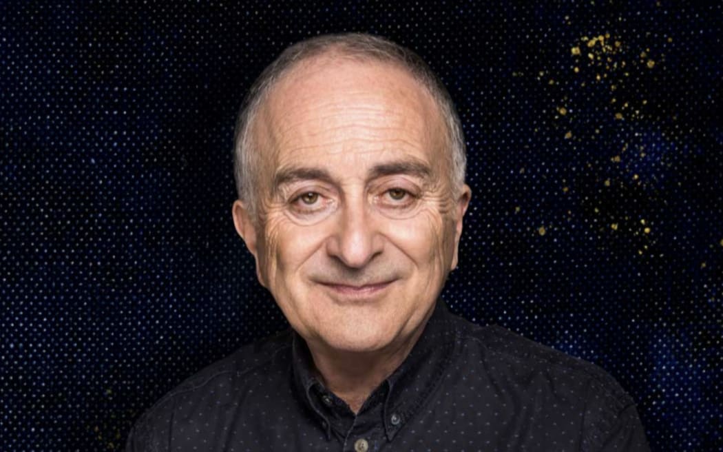 Sir Tony Robinson is a slightly smiling bald man wearing a black shirt with a black background behind him.