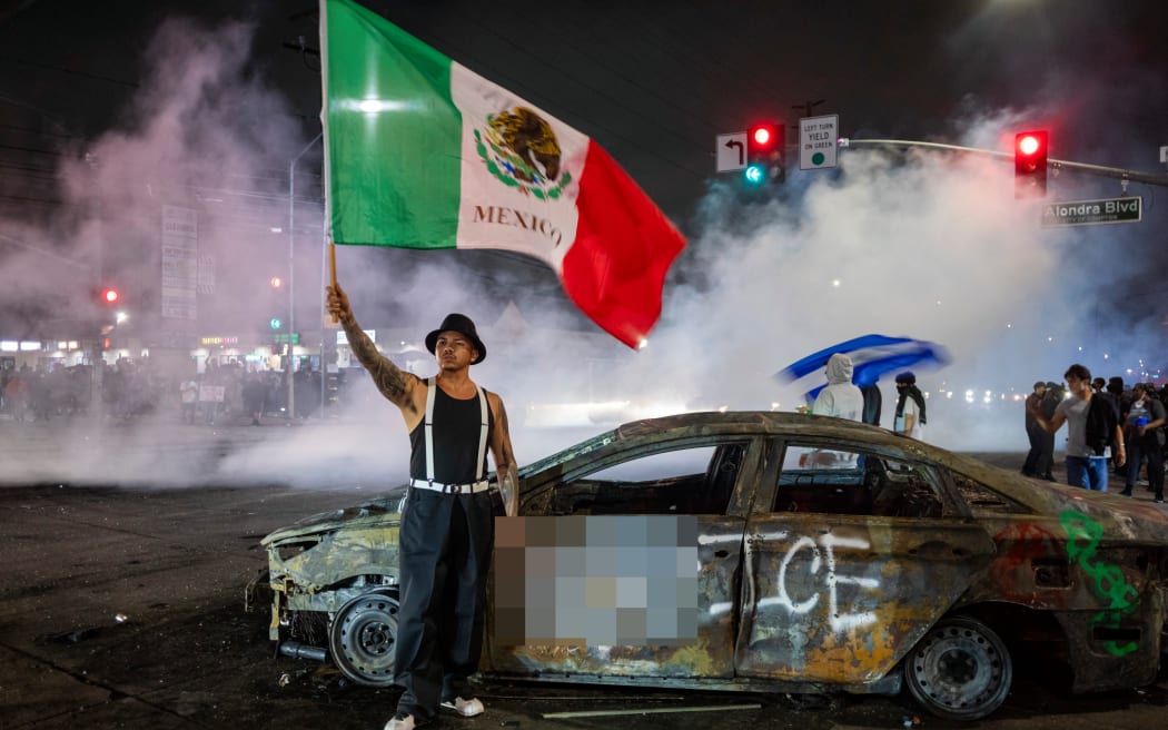 EDITORS NOTE: Graphic content / A protester waves the Mexican flag beside the wreckage of a burnt car sprayed with graffiti against US Immigration and Customs Enforcement (ICE), as law enforcement clashes with demonstrators during a protest following federal immigration operations, in the Compton neighborhood of Los Angeles, California on June 7, 2025. US President Donald Trump deployed 2,000 troops on June 7, 2025 to handle escalating protests against immigration enforcement raids in the Los Angeles area, a move the state's governor termed "purposefully inflammatory." Federal agents clashed with angry crowds in a Los Angeles suburb as protests stretched into a second night Saturday, shooting flash-bang grenades and shutting part of a freeway amid raids on undocumented migrants, reports said. (Photo by RINGO CHIU / AFP)