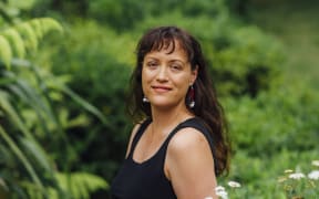 Māori architect Jade Kake wears a black tank top and pants and stands in front of greenery outside.