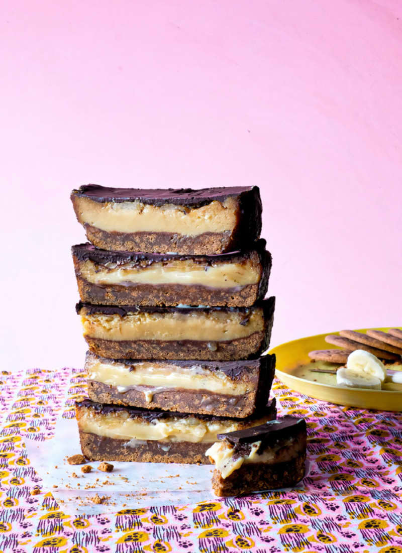 A vertical stack of five pieces of banana caramel slice, showing a cross-section of biscuit base, gooey caramel and chocolate topping, on a pink patterned cloth.