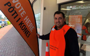 Farrukh smiles at the camera. He is standing outside the entrance to the voting booth at the Pakuranga Plaza, wearing a bright orange vest and holding a large orange voting banner that reads "VOTE HERE - PŌTI I KONEI".