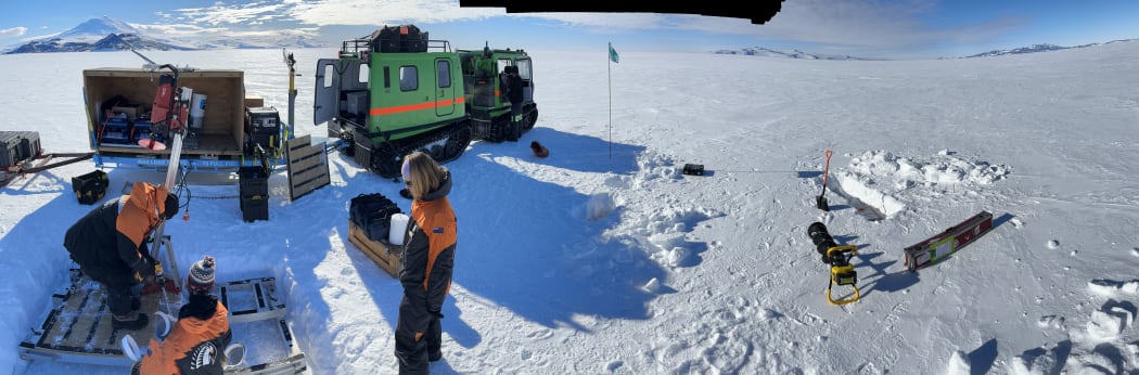 A panorama of a field work site in action on the ice. In the foreground, three people in black-and-orange warm clothing gather around a small platform on the ice, where the drill is being inserted. They are in front of a green hagglund towing a carrier that is open with various gear stacked inside and outside. In the background, streaky clouds in a blue sky above snowy hills.