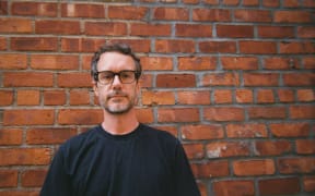 Tony Stamp is a slightly smiling man with glasses and a beard, wearing a black sweater and standing against a brown brick wall.