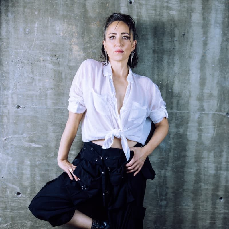 KT Tunstall stands against a wall with short dark hair, a white shirt and a confident pose.