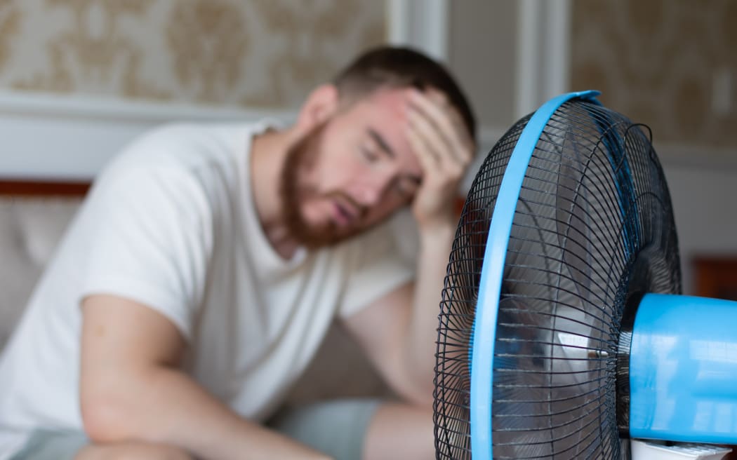 Young bearded man using electric fan at home, sitting on couch cooling off during hot weather, suffering from heat, high temperature