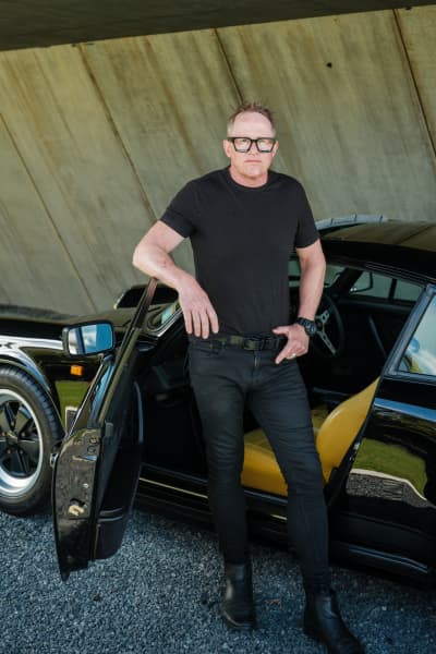 Designer Chris Tate stands with a car door open, underneath Bunker House in Piha.