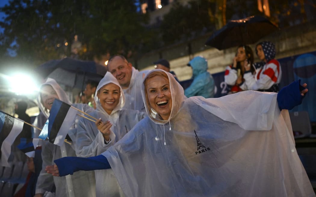Spectators wearing raincoats react as they hold the flags of Estonia along the Seine river embankment during the opening ceremony of the Paris 2024 Olympic Games in Paris on July 26, 2024. (Photo by Natalia KOLESNIKOVA / AFP)