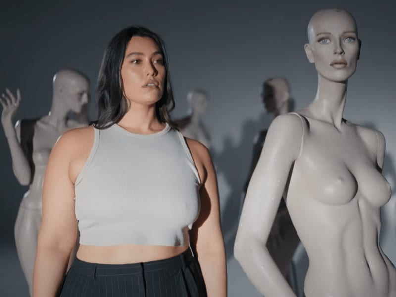 Isabella Moore - a woman with long brown hair and a grey tank top - stands among a group of thin mannequins.