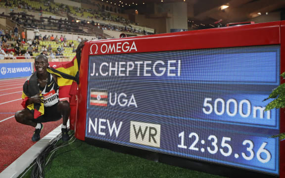 Uganda's Joshua Cheptegei, wearing an Uganda national flag on his shoulders, poses for pictures next to the timer screen after breaking the world record in the men's 5000metre event during the Diamond League Athletics Meeting at The Louis II Stadium in Monaco on August 14, 2020.