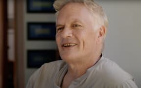 A smiling white-haired man in a grey t-shirt looks to the right of camera.