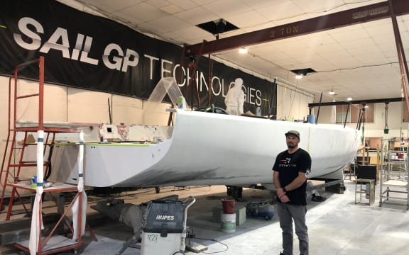 Malcolm stands next to a large, white, metal structure. It's held up on a platform and still under construction. In the background is a sign saying "SAILGP TECHNOLOGIES"