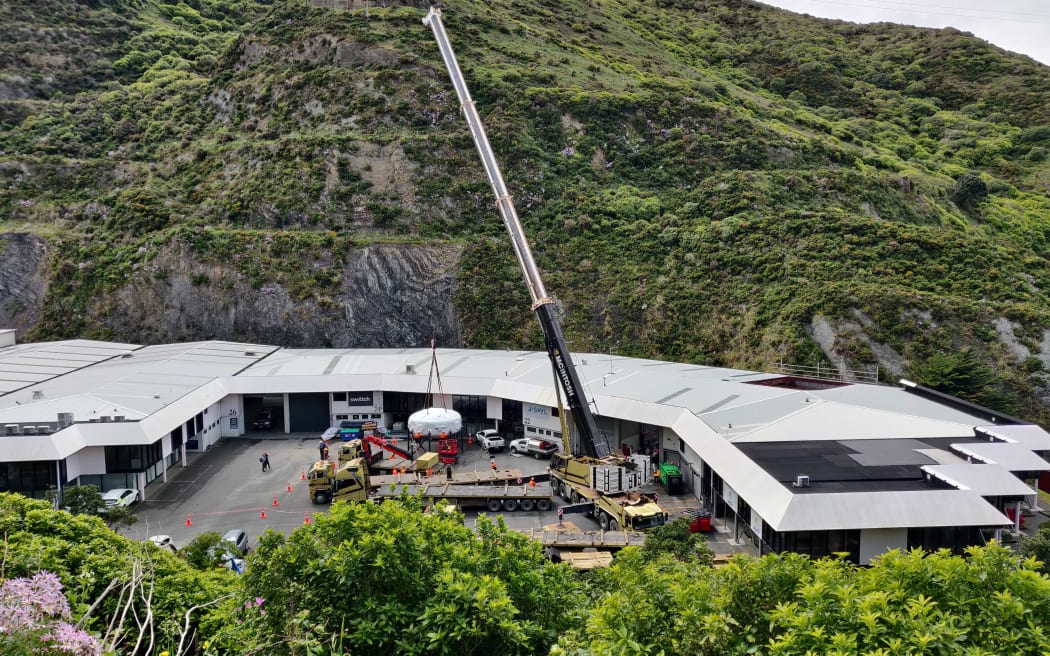 Viewed from a high vantage point, a tall crane lowers a car-sized white flat cylinder - shaped like a bagel without the hole - in the carpark next to a single-storey building framed by steep green hillsides.