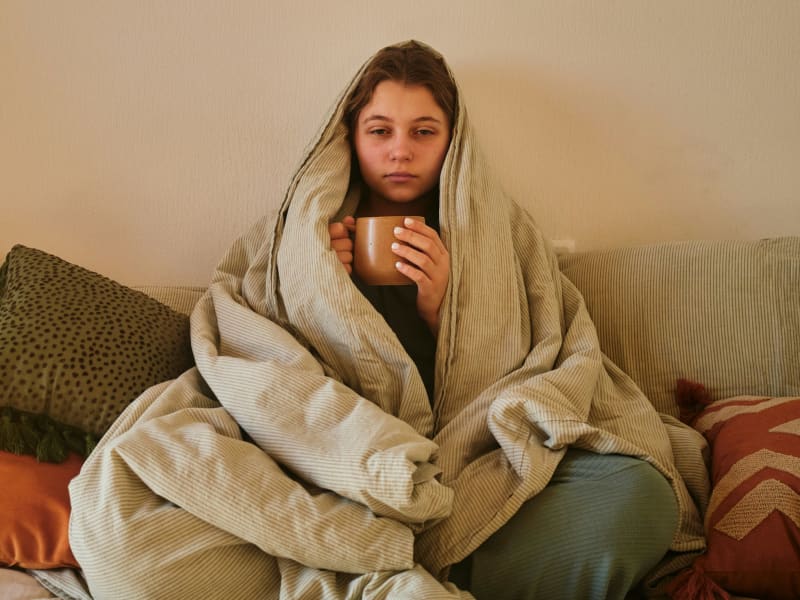 Woman looking tired and sick, wrapped up in blanket on couch, holding a mug.