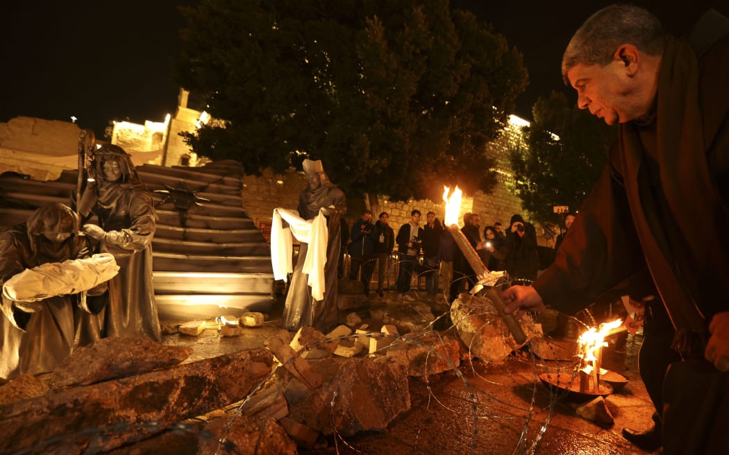 A priest places a candle in front of an installation depicting the nativity of Christ with a figure "symbolizing baby Jesus lying in his manger amid rubble", in reference to Gaza, is displayed in front of the Church of the Nativity in the city of Bethlehem in the occupied West Bank on December 23, 2023. (Photo by HAZEM BADER / AFP)