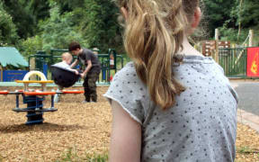 A photo of a lonely girl at the playground watching other kids playing.
