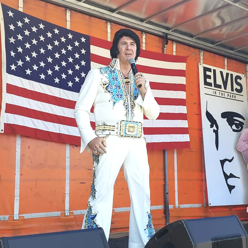 An Elvis impersonator stands on a stage on the back of a truck with an American flag stretched out on the wall behind him.