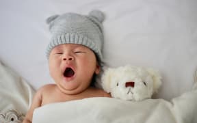 Image of a yawning baby wearing a knitted grey beanie hat tucked into cot with white teddy bear poking above the sheet.