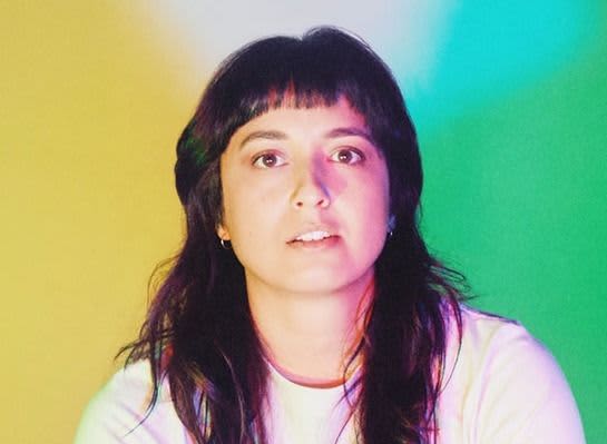 Liz Stokes - a young woman with long brown hair - gazes at the camera with a colourful background behind her.