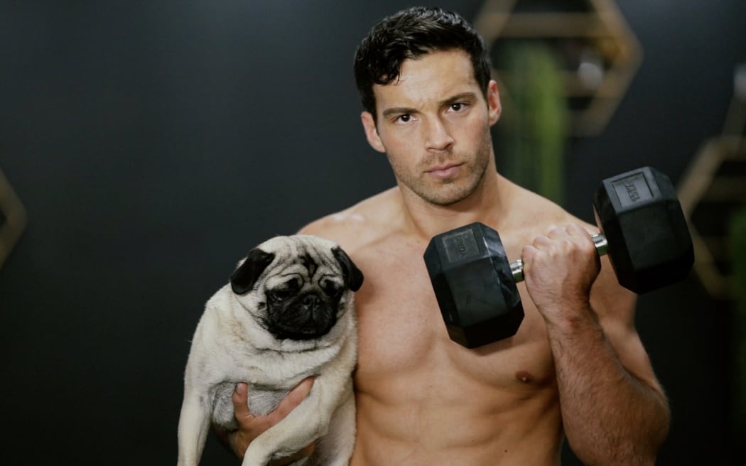Shirtless social media influencer and fitness coach Daniel Rankin poses with a handweight and a small grey pug.