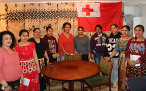 Standing in front of a large tapa cloth and a Tongan flag, the nine Tongan women of the night shift gather to support each other.