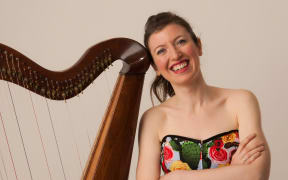 Una leans next to her harp. She is wearing a brightly-coloured dress and smiling.