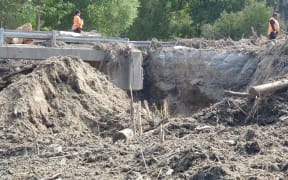 Roadworkers stand either side of a 2m wide, 3m deep gash across the width of SH5 adjacent to Marshall's Bridge near Glengarry. A crossing to the side allows contractors' vehicles across.