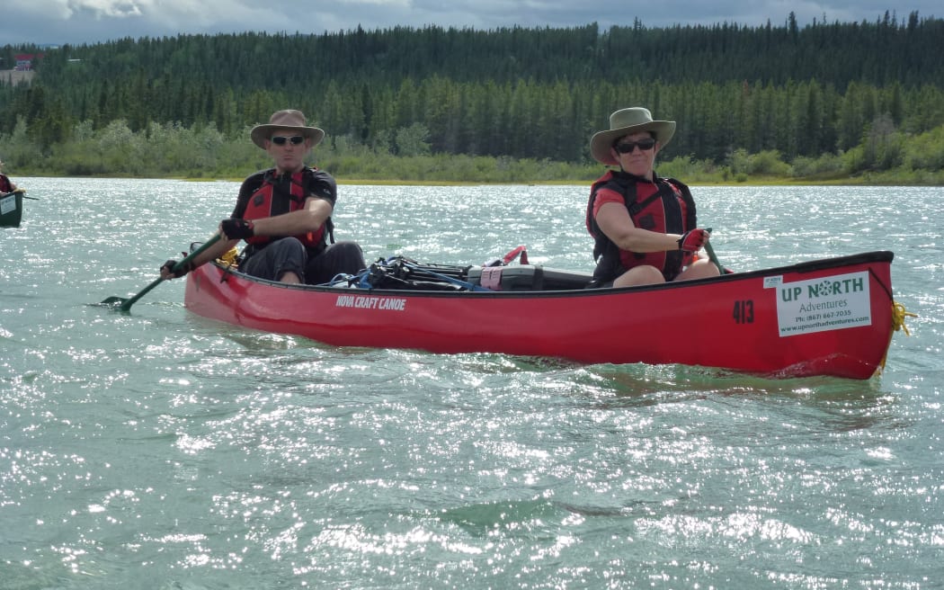 Two people sit in a red kayak on the Yukon river. They are both wearing lifejackets and hats. There is a forest on the bank behind them.
