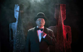 Image from Tame Iti: I Will Not Speak Māori Exhibition.  Artist wearing a suit, bowtie and bowler hat stands in front of metal sculptures in a stylised human form.