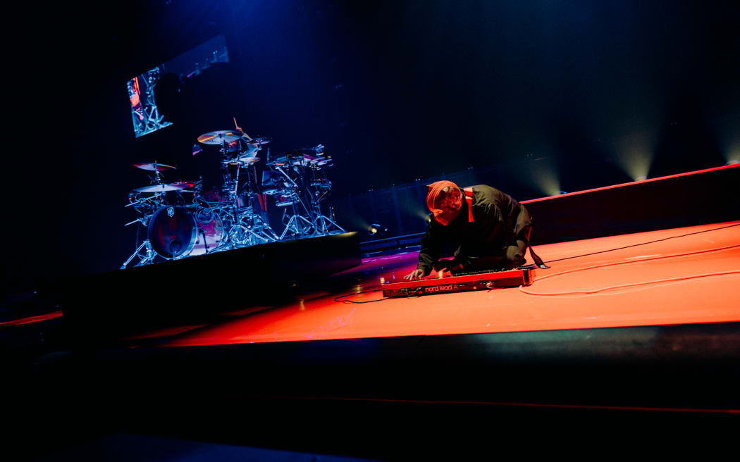 A man crouching on the ground in front of a synth with a man playing drums in the background