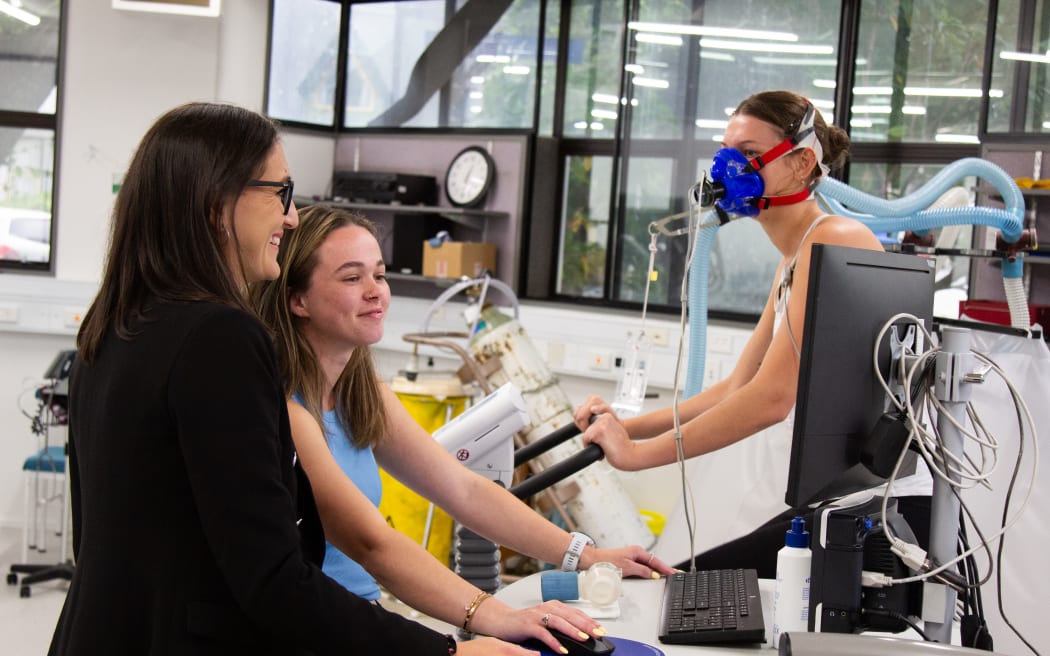 Two researchers are at a desk with a computer, looking at the monitor, while a research participant is on an exercise bike behind them, with a mask on that has tubes coming from it.