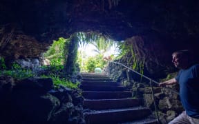 A wide shot of a man standing in a cave at the bottom of stairs that head up towards a light and leafy backyard.