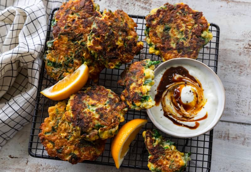 A wire tray topped with corn, green chilli, spinach and cheese fritters, garnished with lemon wedges and a bowl containing chutney swirled through yoghurt.