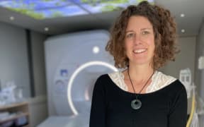A woman with a curly bob, wearing a white lace top underneath a black jersey and a pounamu necklace, smiles at the camera. She is standing in front of an MRI machine.