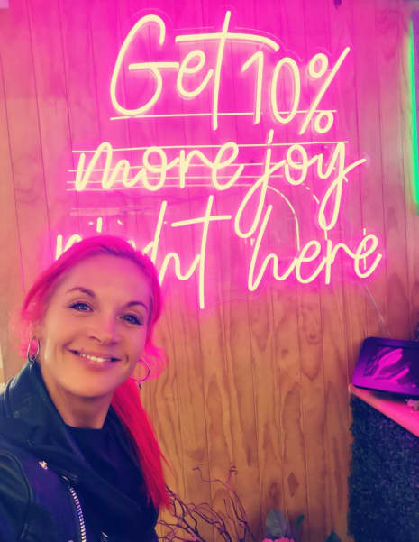 Mindset and behaviour change coach Bex Bell smiles next to a sign that reads 'get 10 percent more joy right here'.