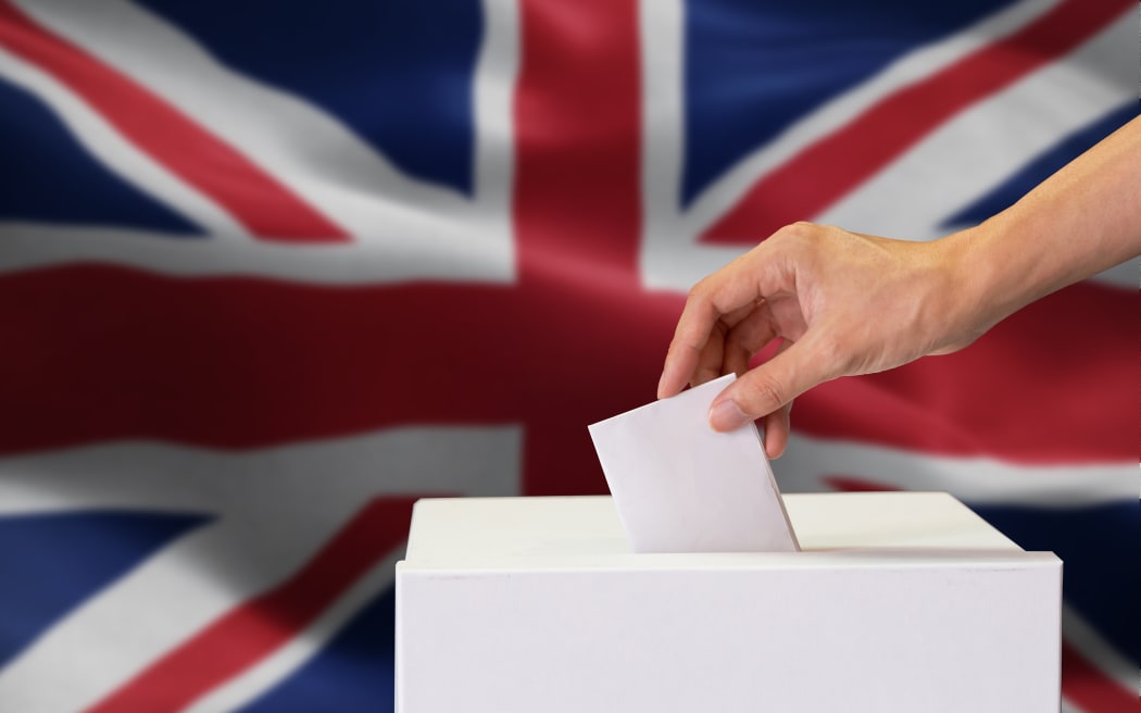 Close-up of man casting and inserting a vote and choosing and making a decision what he wants in polling box with United Kingdom or UK flag blended in background.