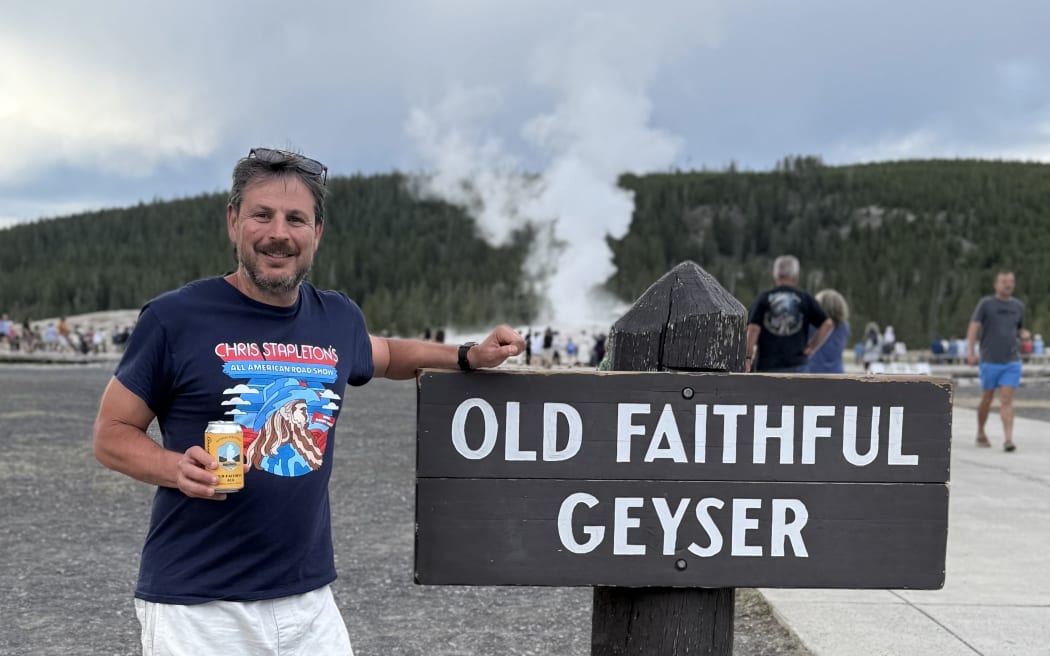 Ash Jurberg is holding a can of Yellowstone branded beer and standing next to a sign that says "Old Faithful Geyser". In the background you can see steam coming off the geyser.