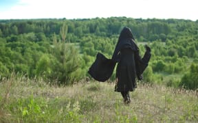 woman witch in a black cloak looks into the woods from a mountain covered with grass