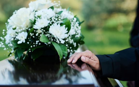 Focus on hand of mourning mature woman in black attire on lid of closed wooden coffin with bunch of fresh white chrysanthemums on top