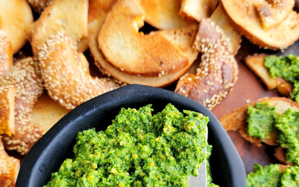 This is an image of a small bowl of bright green rocket pesto next to some toasted bagel halves.