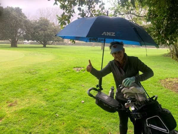Psychologist Karen Nimmo gives a thumbs up of herself while standing with her golf bag gear under an umbrella.