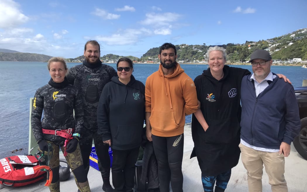 Six people stand in front of the ocean underneath a blue sky with the hills of Wellington in the background. The two people on the left are wearing wetsuits.