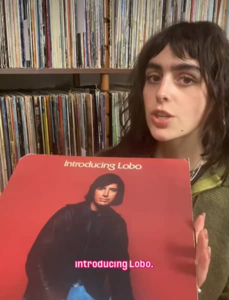A young woman wearing a green top holds up a record titled 'Introducing Lobo', against a backdrop of shelves full of records.