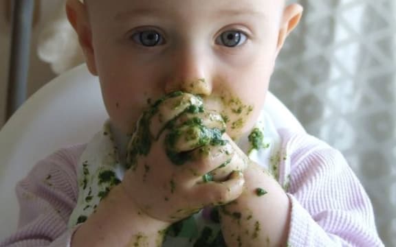 A photo of a toddler eating messy green food with her fingers