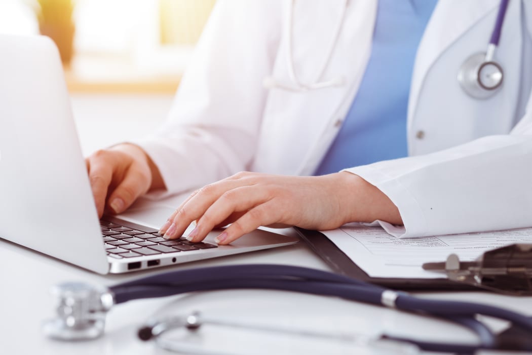 Unknown woman-doctor typing on laptop computer while sitting at the table in sunny clinic.
