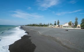 A view along the gray stony Pacific Beach with walkway, Marine Parade Gardens and the soundshell, with Napier city buildings in background.
